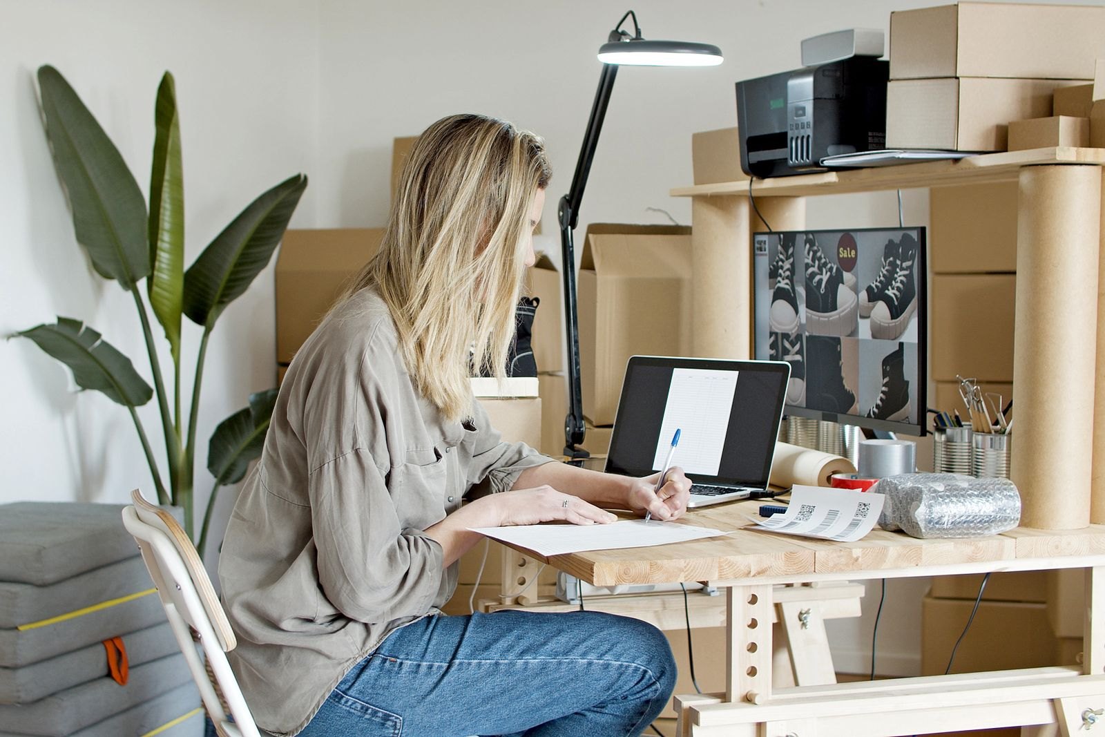 A woman writes on paperwork beside a laptop and packing supplies at a worktable.