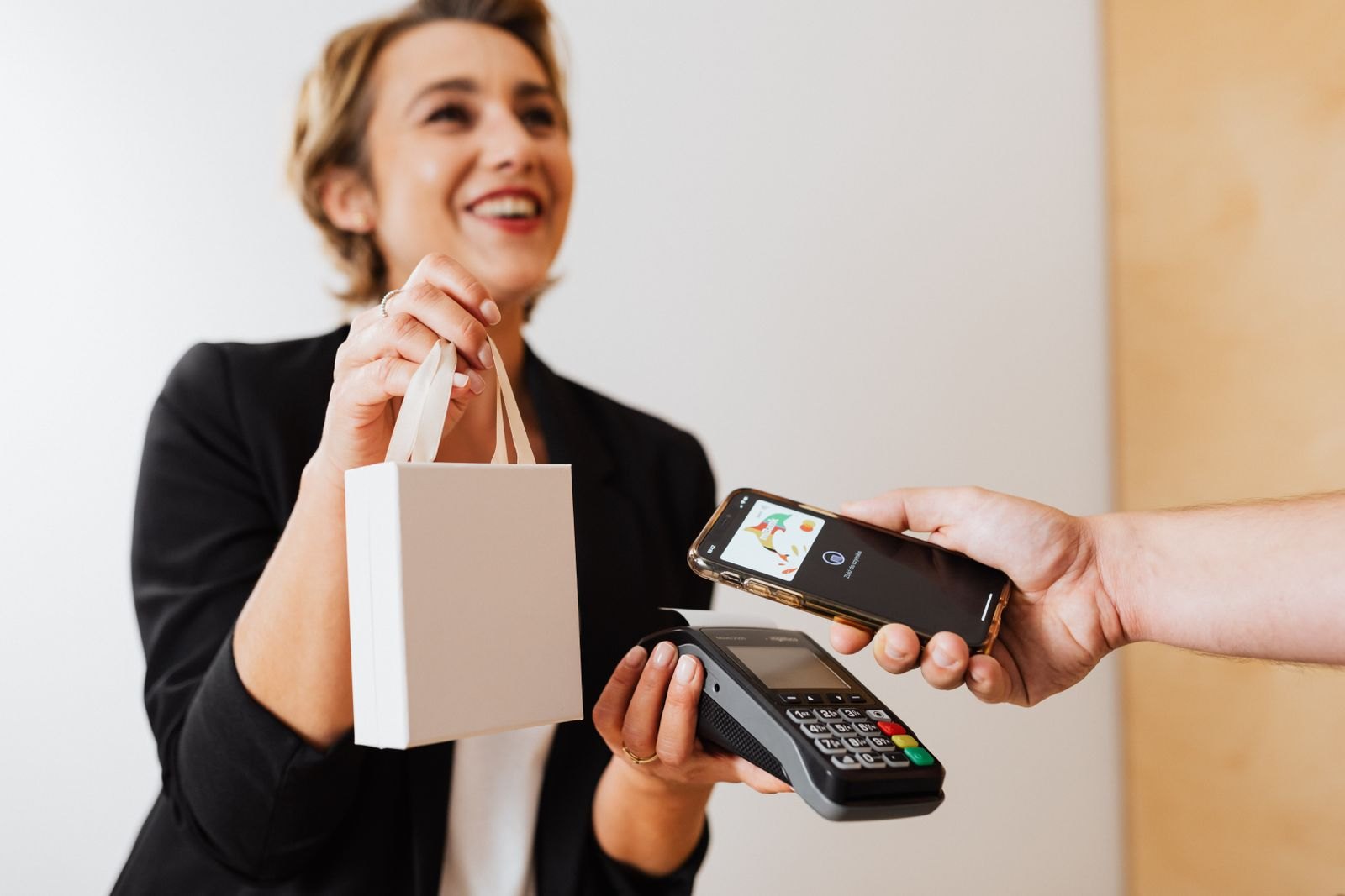 A customer holds a smartphone over a card terminal while a shop assistant holds a shopping bag.
