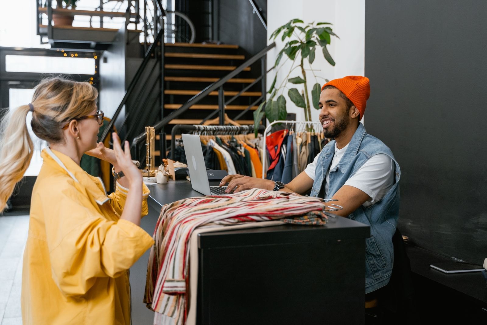 A clothing shop assistant and a customer talk across a counter beside a laptop.