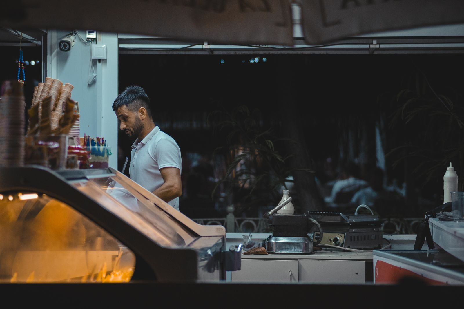 A worker behind an ice cream shop counter with cones and serving equipment.